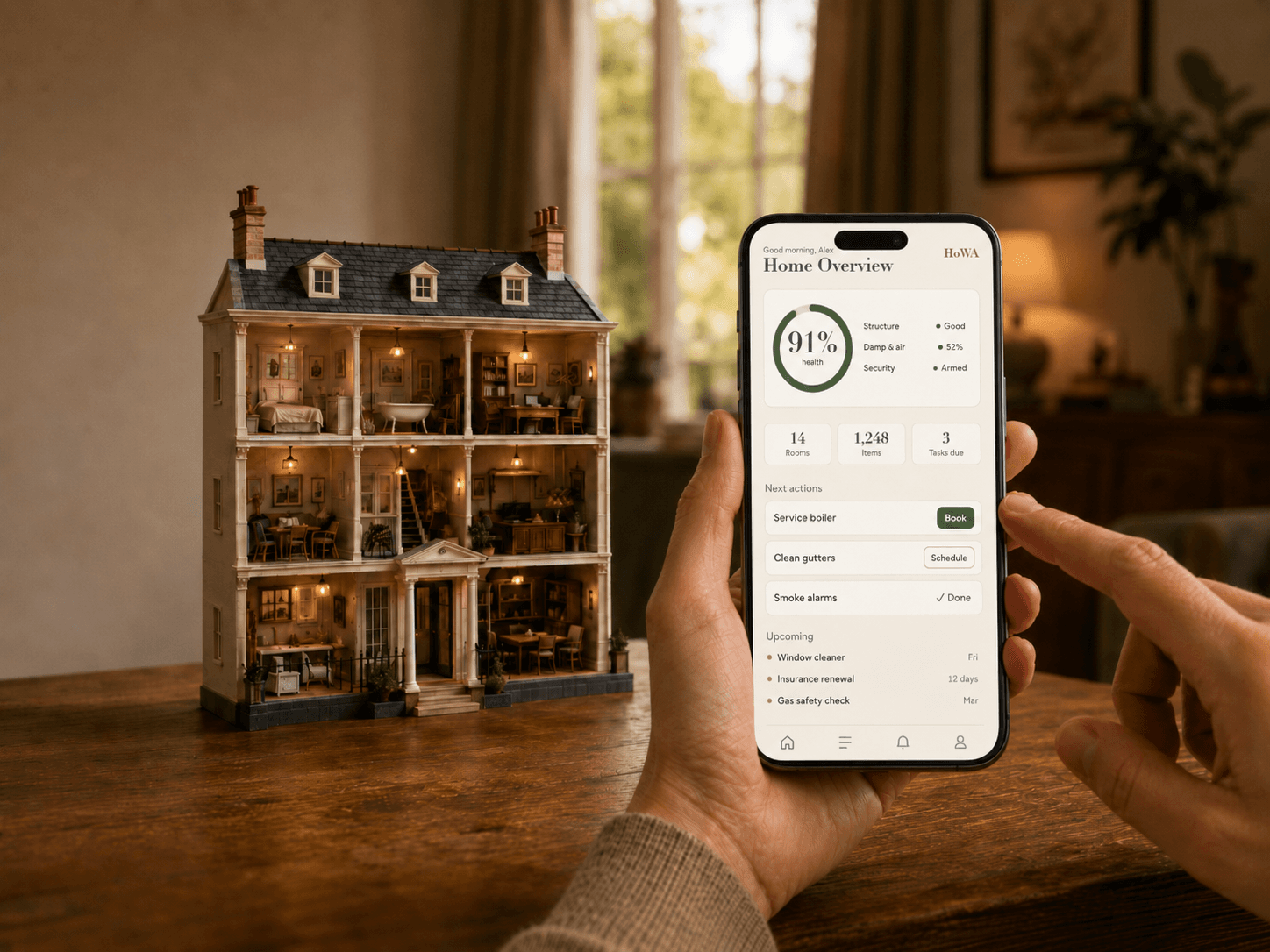 A person holding a phone showing the HoWA home dashboard beside a cutaway dollhouse on a table.